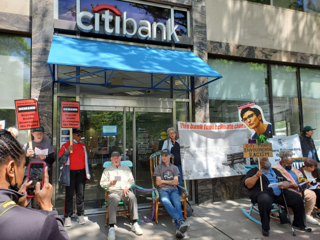 Older adults sitting in rocking chairs and standing holding signs blocking the entrance of a Citibank branch. Signs read, This bank funds climate chaos, and Demand, boycott banks destroying the planet.