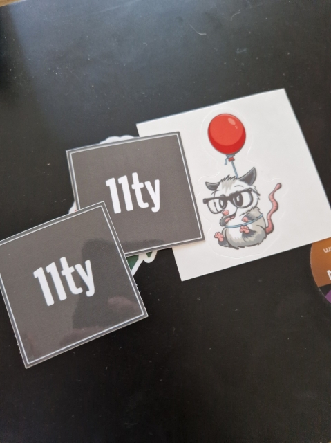 Photo of two 11ty stickers and a possum dangling from a balloon, wearing glasses and a hula hoop.