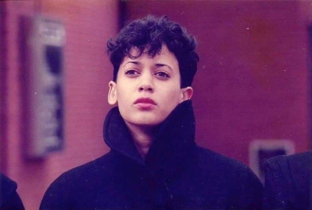 A young Kamala Harris, wearing a black coat with a high collar in front of a blurred maroon background. She has short hair and kind of a wistful look on her unsmiling gorgeous face. She about to drop the most fire synth album ever.