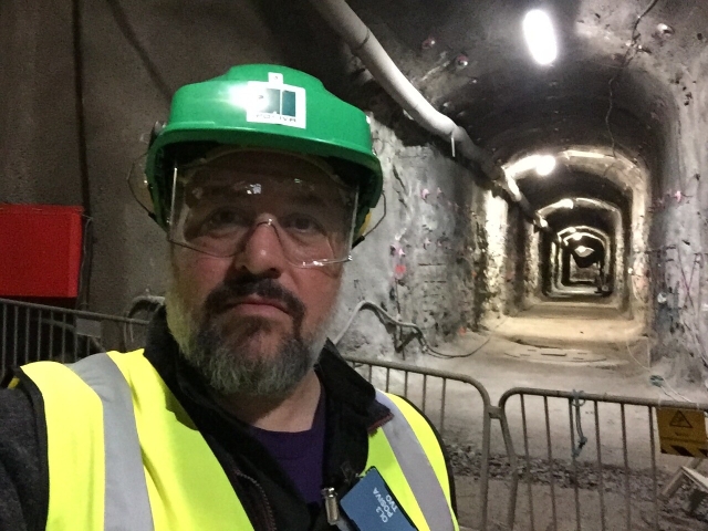 Image of a man with a green hard hat standing in an underground tunnel. Me in Onkalo in 2016.