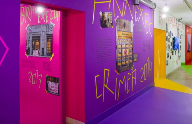 The Haus De Kunst Museum exhibition showing sections of bright purple, white and red walls interrupted by bright pink and yellow doorways leading into other rooms. Lettering made from yellow tape reads: Annexed Crimea 2014, along with photographs and other images also secured to the walls in the same adhesive tape.

Included in the display are handwritten texts, videos and photographs.
