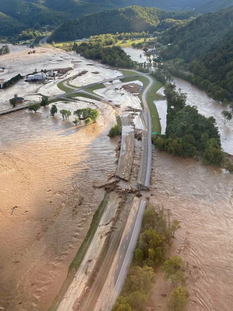 A high view over a hilly mountainous green landscape with a highway that crosses an opaque reddish river which dominates the foreground, but the river is swollen and has broken the bridge so it is impassable. The water also fills every hollow of the hills and comes partway up many trees.