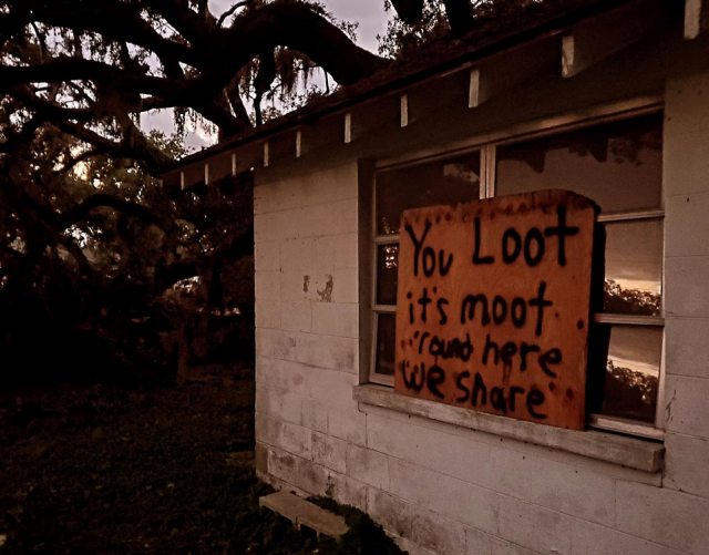 Wooden sign on side of home over widows reading in black paint, "You Loot, it's moot, round here, we share." 