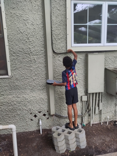 A child in a red and blue striped soccer Jersey with black shorts and black crocs stands on some cement blocks with a screwdriver in his right hand, unscrewing something from a window frame. 