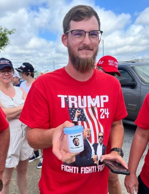 A Trump supporter holding a plastic specimen cup containing their sperm that has a picture of JD Vance on it with the words:

"JD Vance: Full Family Kit" on it.