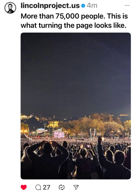 A large crowd gathers at an outdoor event at night, with a stage visible in the distance and people raising their hands in the foreground.