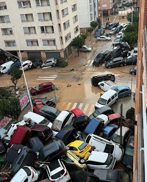 A chaotic scene of overturned and damaged vehicles piled against buildings on a flooded street, with water pooling around the cars and debris scattered throughout the area.