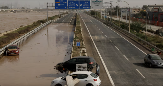 After the extreme rain events this is a picture from the El País newspaper showing a stretch of the V-30 that runs alongside the Turia river. The river is in full flood, very wide with brown turbulent water on the left. In the centre of the picture, the road is partially flooded and there are cars abandoned and debris overtop the road.  A roadway sign stretches over the 4 lanes of traffic.