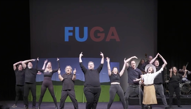 The cast of "Young Greg Abbott: A FuQusical" line up on stage, middle fingers raised. They are dressed in simple, all-black clothing except for the band leader, who is in Western wear: a tan skirt, pale colored Western button down, and a cowboy hat. Behind them to the right is the band on a small riser, and on a screen behind the cast is the acronym FUGA: Fuck You, Greg Abbott.