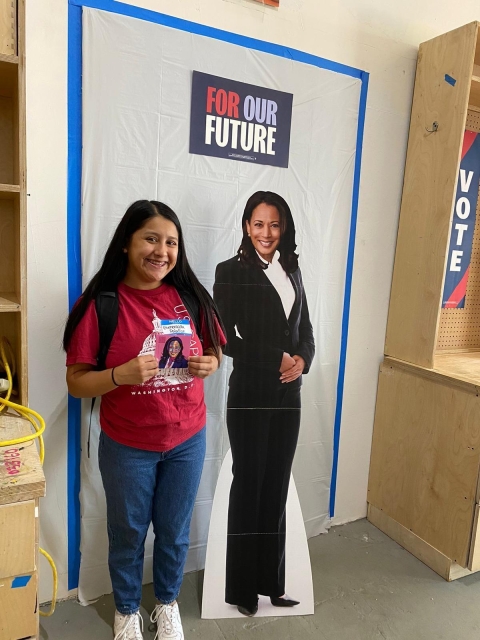 A young Latina girl standing next to a Kamala Harris figure with a for or future sign behind her and a vote sign to the right of her. She is wearing a red shirt with the capital on it saying Washington DC holding up a picture of Kamala Harris