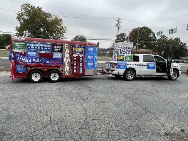 A truck is towing a trailer decorated with political campaign signs supporting Kamala Harris and Tim Walz for the 2024 election. The truck has an open door, and various messages promoting voting and regulating gun laws are displayed on both the truck and trailer