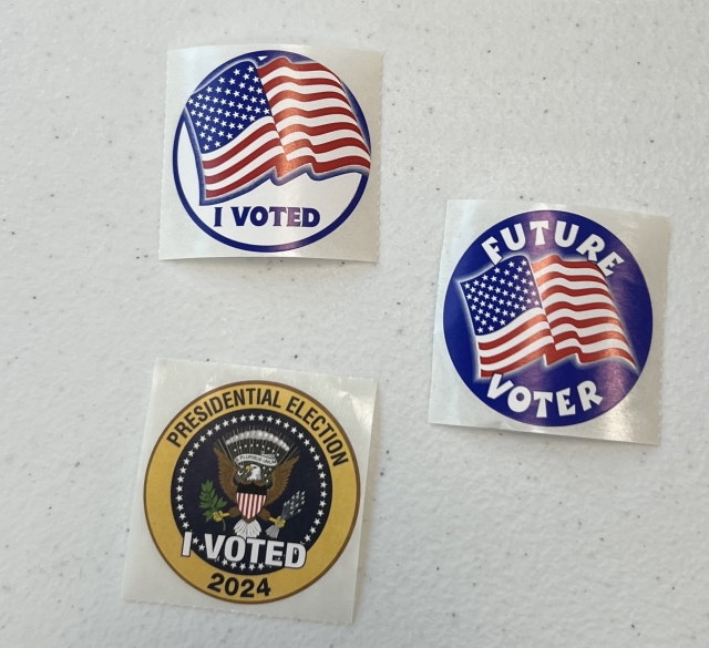 Photo of 3 stickers. One is the American flag with a simple “i voted”. One is a similar design of a waving flag, but it’s on a dark blue background and it says “Future Voter”. The third is based on the seal of the President of the US and it says “I Voted” in the middle and “presidential election 2024” around the edge. 