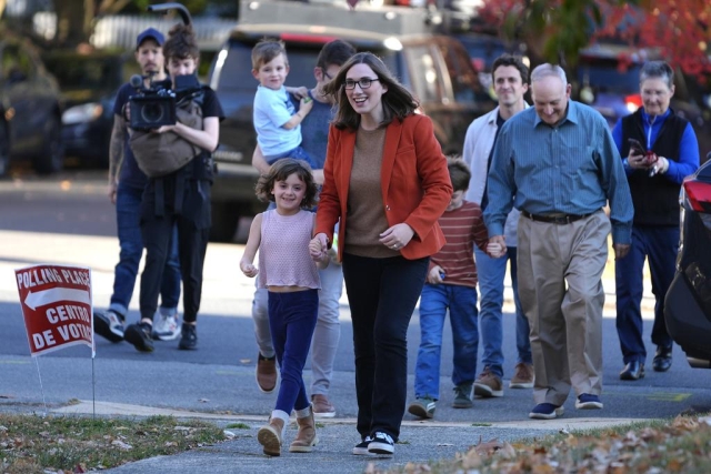 A woman wearing glasses and a red blazer walks down a sidewalk toward the camera, holding the hand of a child. 