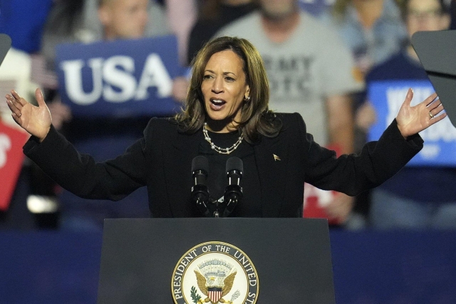 Kamala Harris stands in front of a podium, arms outstretched. Behind her is a crowd of supporters. 