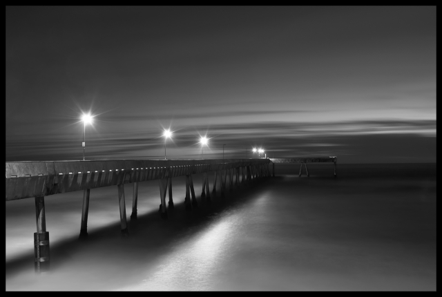 It's a half hour past sunset. The sky has a slight bit of light remaining. There are a few streaky clouds near the horizon... nothing threatening, just wisps.

Looking west across the ocean, a tall pier with stilt-like concrete legs reaches out above very calm waters. A few starry streetlights rise above the pier, castng glowy light onto the smooth water below.