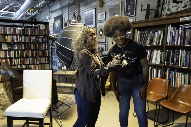 Visual fellow Sarahbeth Maney shows a Black community member her camera. Behind them are bookcases and chairs.