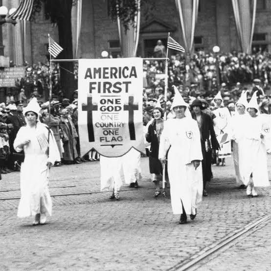 Picture of KKK people walking in their sheets with a banner that reads America first at some kind of a parade. It looks like.