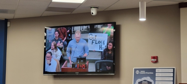 A speaker on the council livestream is shown on a TV on the wall. Behind them a person holds a sign that reads "Get the Flock outta here"