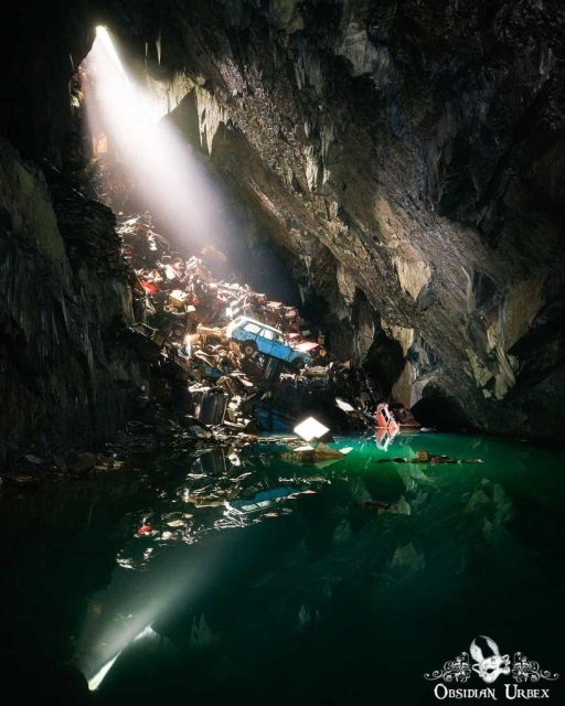 A sunlit cave containing a body of green water is filled with illegally dumped cars