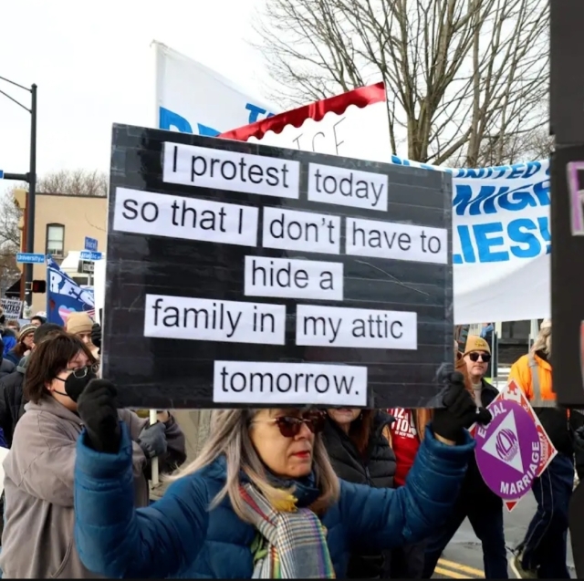 A woman  carrying a protest sign that reads: I protest today so that I don't have to hide a family in my attic tomorrow 