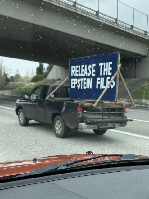 Photo of a small black pickup truck in the right lane of a highway about to go under an overpass. In the back of the truck is a homemade billboard on a 4 by 8 foot piece of plywood. The background is dark blue and I large white block print it reads:
RELEASE THE
EPSTEIN FILES.