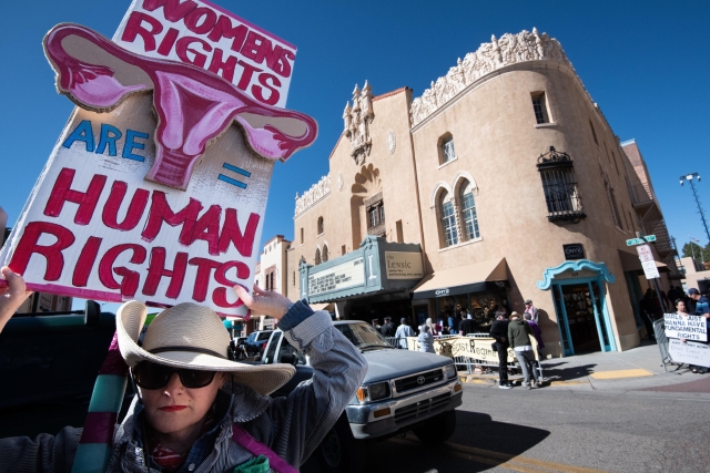 Woman in a black and white hat holding a large sign reading Women’s Rights Are Human Rights, in front of the historic Lensic Theater. The sign includes a prominent illustration of female reproductive organs. 