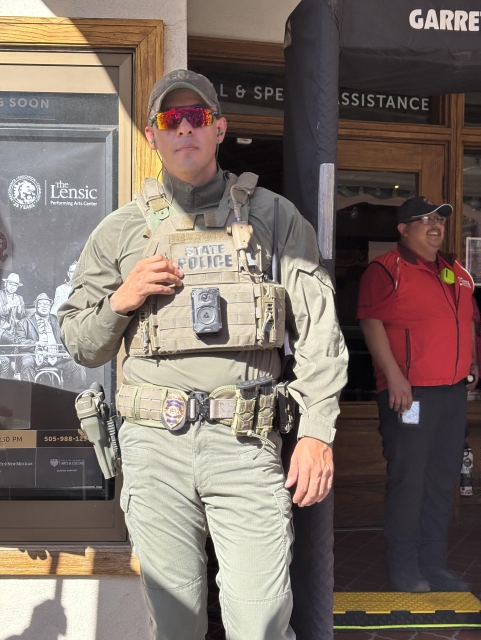Member of New Mexico State Police in camo and weaponry standing in front of the Lensic Theater entrance, where metal detectors are set up to screen attendees. In the background is a smiling member of a private security team. 