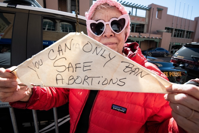 Woman in pink hat and pink heart-shaped sunglasses holds a hanger. Paper on it reads “you can only ban SAFE abortions”.