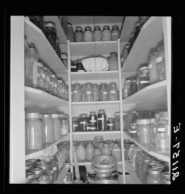 The image depicts a well-organized pantry filled with numerous jars of preserved food. The shelves are neatly arranged, showcasing an assortment of items such as pickles and jams in clear glass jars. Labels on some of the jars indicate different contents or possibly production dates. This storage space is indicative of careful preservation practices, likely aimed at extending the shelf life of perishable goods for consumption during times when fresh produce might be unavailable.

The black-and-white nature of the photograph suggests it could be from a past era, reflecting traditional methods of food conservation before modern refrigeration techniques became widespread. The organized arrangement and variety of preserved foods highlight an approach to self-sufficiency in sustenance that is both practical and resourceful.
